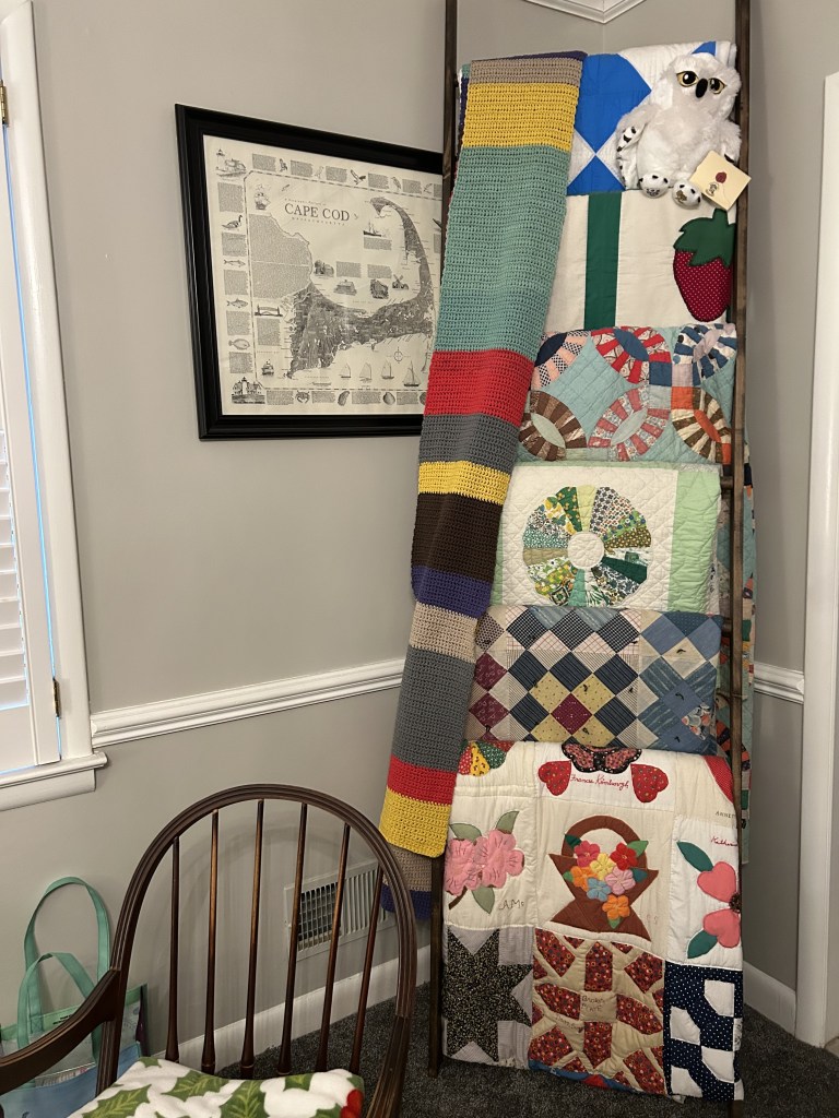 Image of a quilt ladder displayed in a cozy room. The ladder holds a collection of colorful handmade quilts, including a double wedding band quilt, a basket design, and others with intricate patterns. A crocheted striped blanket hangs alongside them. At the top of the ladder is a plush white owl. A framed Cape Cod map hangs on the wall, with a wooden chair and a tote bag visible in the foreground.