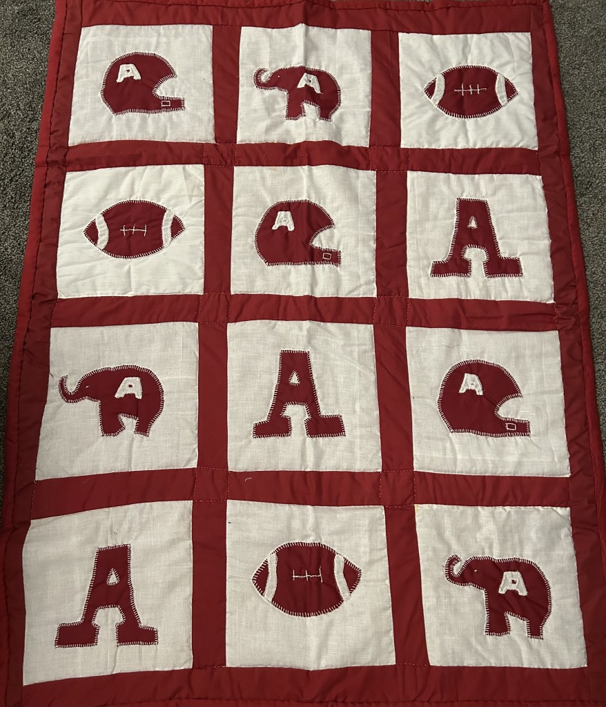 Image of a handmade quilt with a crimson and white Alabama football theme. The quilt features appliqué designs of football helmets, footballs, the letter 'A,' and elephants in alternating squares. Each design is outlined with visible stitching, and the quilt is bordered with a crimson edge, showcasing school spirit and craftsmanship.